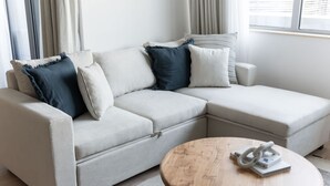 En moderne stue med en beige hjørnesofa, et rundt træsofabord og et vindue med gardiner.