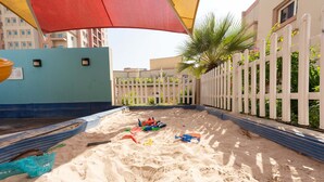 A sandy play area with toys, a blue wall, a red umbrella, and a white fence. - Spacious Studio at Imperial Residence in JVT That Sleeps 4 (Dubai)
