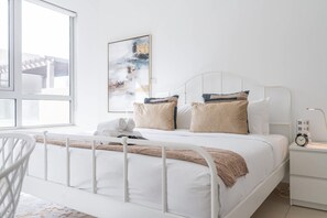 Ein ordentlich bezogenes Bett mit weißem Metallgestell, beigen und blauen Kissen und einer braunen Tagesdecke. Ein weißer Nachttisch mit einer Uhr und einer Steckdose ist sichtbar.