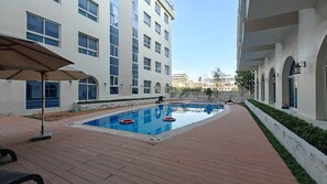 A swimming pool area with a clear view of the pool, a lounge chair, and a building with multiple windows.
