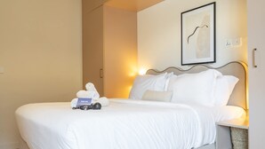 Ein ordentlich gemachtes Bett mit weißer Bettwäsche, ein gerahmtes Kunstwerk an der Wand und ein Nachttisch mit einer Lampe und einem kleinen Korb.