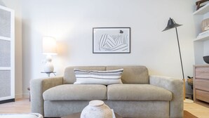 Ein Wohnzimmer mit einem beigefarbenen Sofa, einer weißen Vase auf einem Couchtisch, einer Stehlampe und einem gerahmten abstrakten Kunstwerk an der Wand.