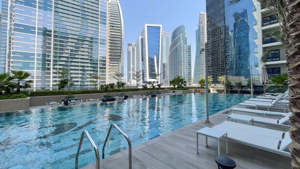 A swimming pool with lounge chairs and a palm tree, surrounded by high-rise buildings. - Excellent 1 Bedroom in Zada Tower With Balcony (Dubai)