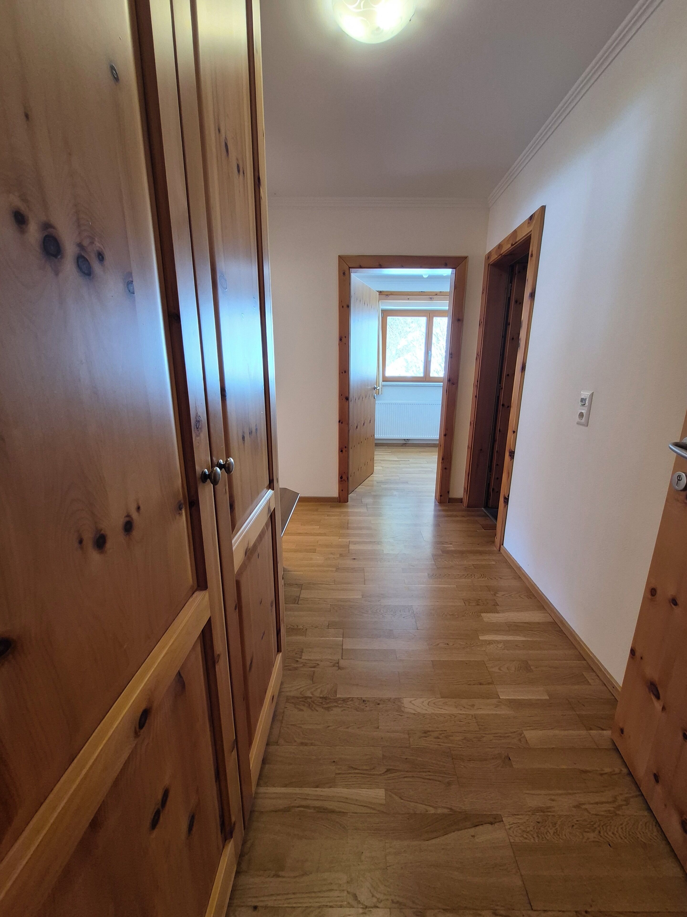 Un pasillo con suelo de madera y puertas, que conduce a una habitación con ventana.