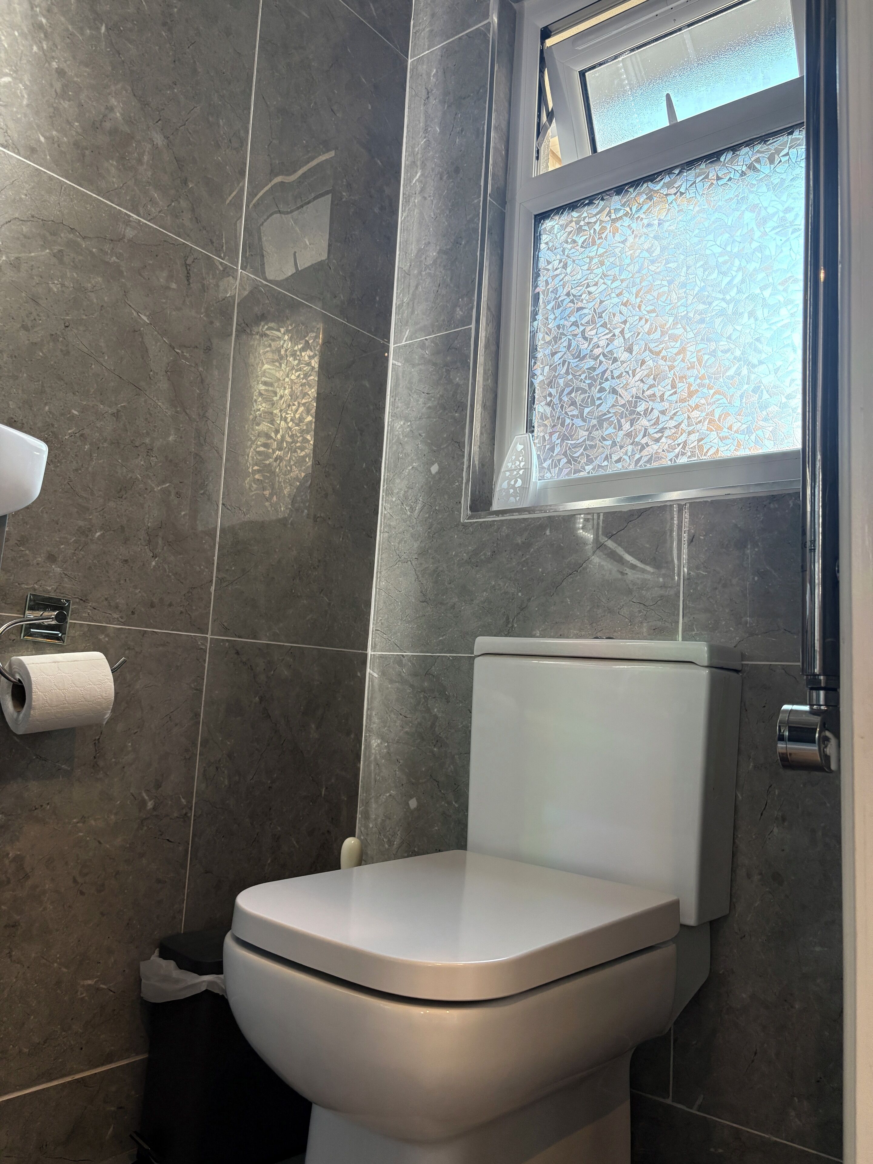 A bathroom with a toilet, a paper towel dispenser, and a window with a patterned glass.