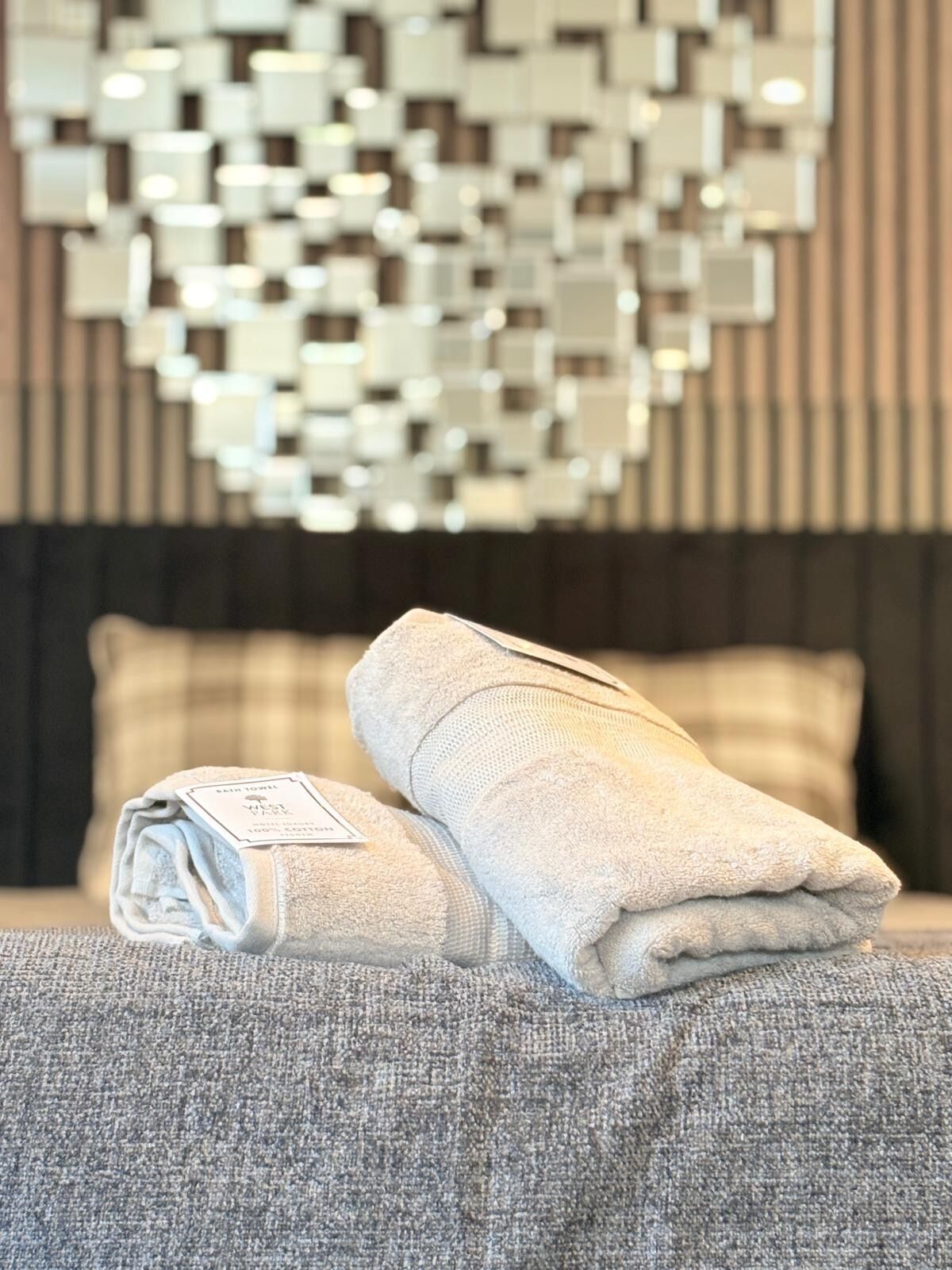 A neatly made bed with a folded towel and a tag, a textured gray pillow, and a wall with a geometric mirror design.