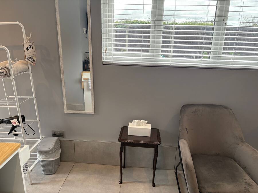 Une pièce avec un mur gris, une fenêtre avec des stores, un miroir, une petite table avec une boîte de mouchoirs, une chaise grise, une étagère blanche avec une serviette et une poubelle.