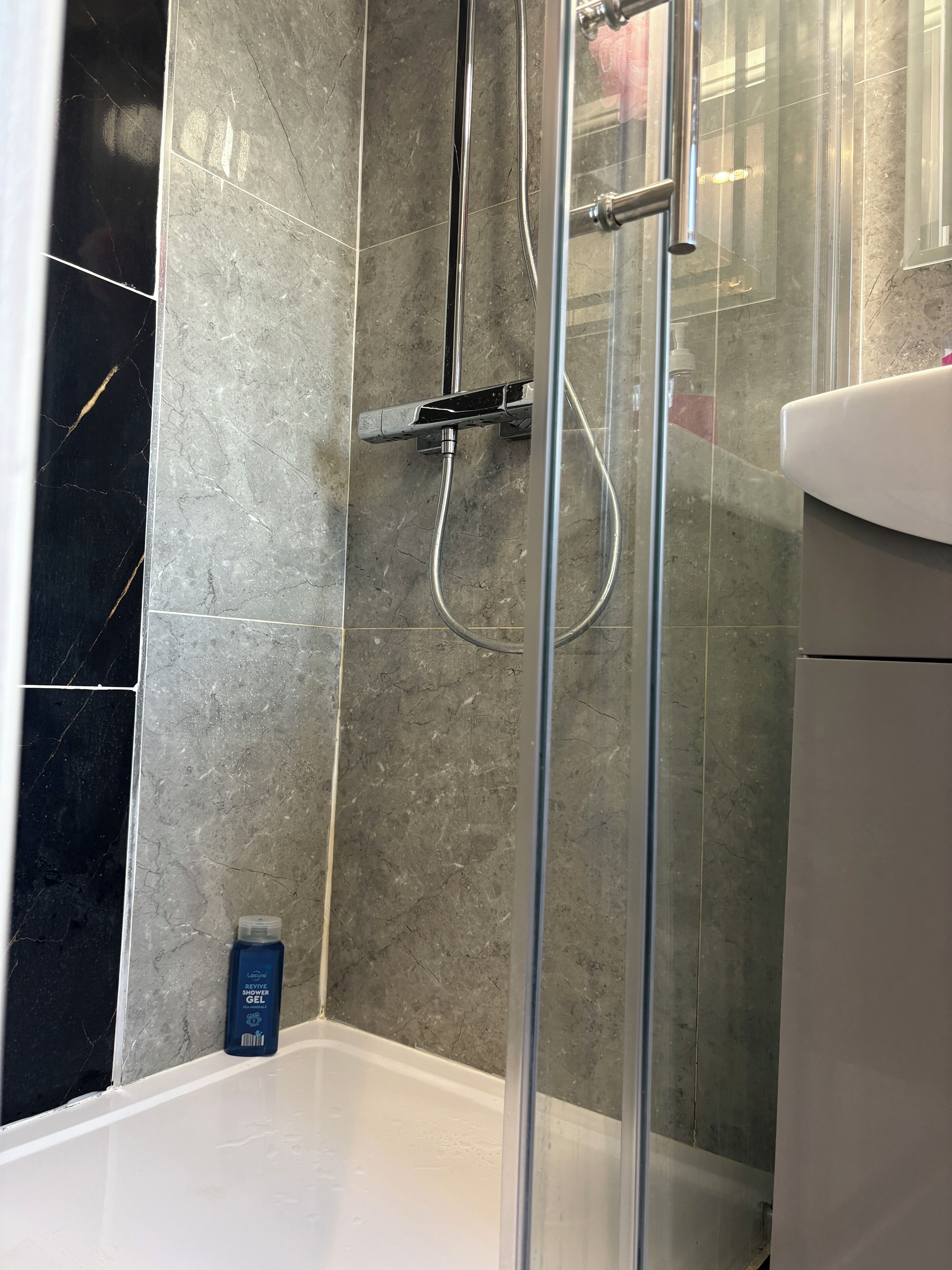 A bathroom with a glass-enclosed shower, a white bathtub, and a bottle of shampoo on the floor.