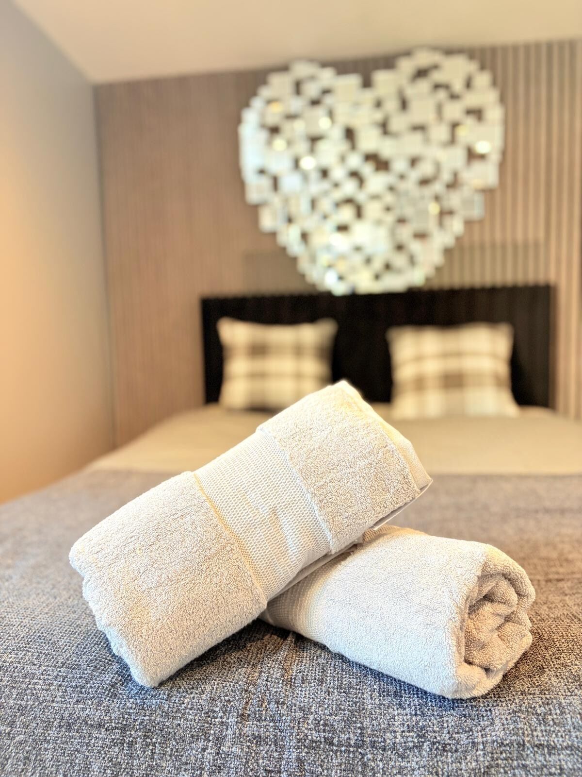 Un lit bien fait, agrémenté d’une décoration murale en forme de cœur et de deux serviettes enroulées posées sur un couvre-lit gris texturé.