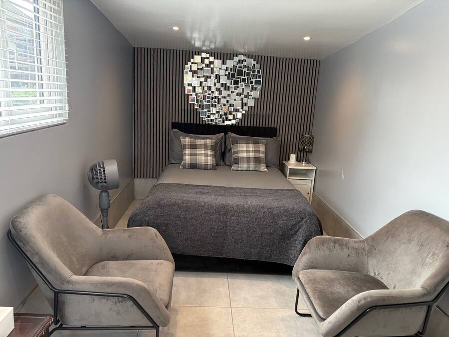 A modern bedroom with a large bed, two armchairs, a wall art piece, and a bedside table with a lamp.