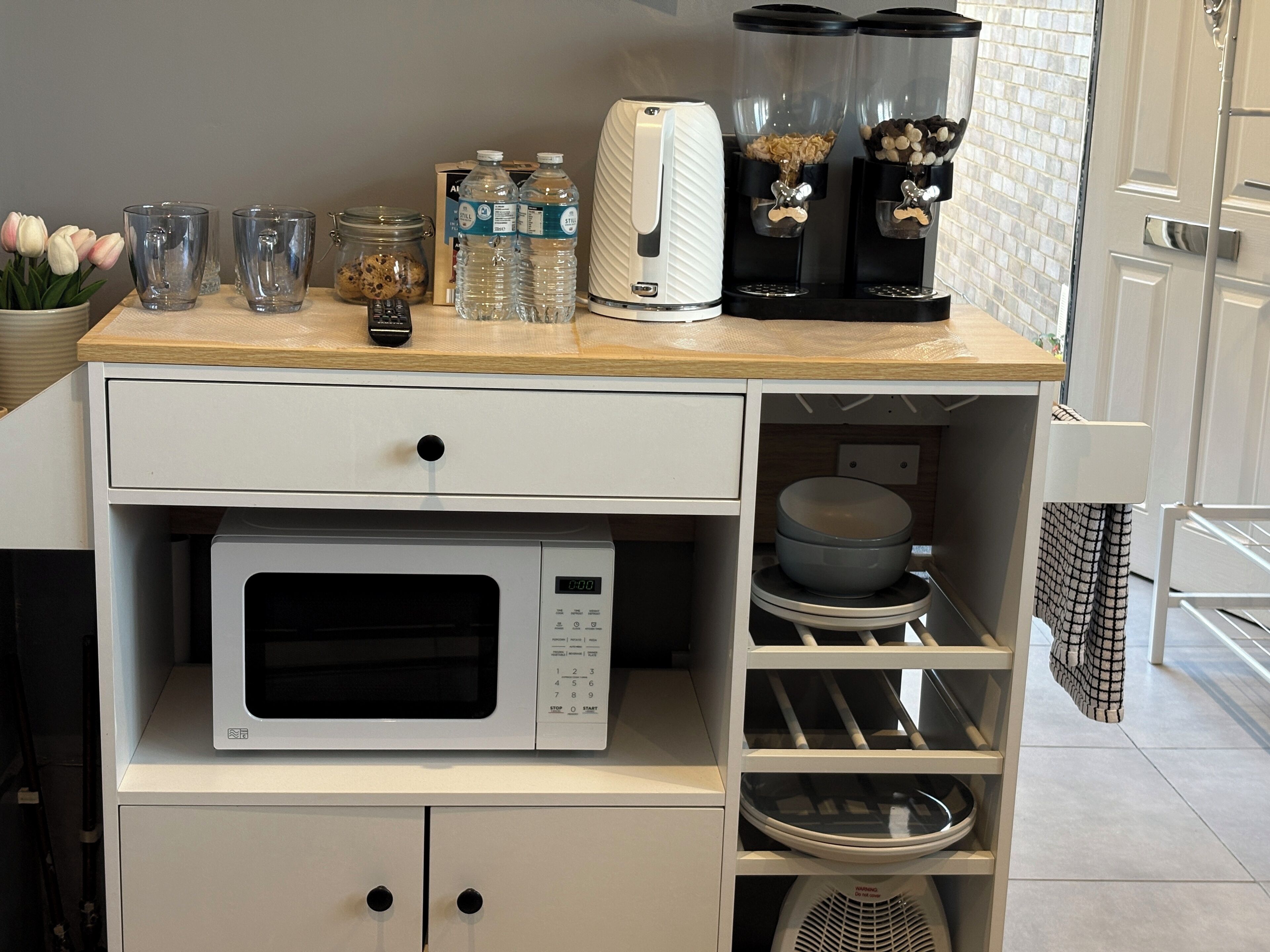Una cocina con microondas, tostadora y cafetera sobre una encimera de madera. Hay frascos y un bol en los estantes inferiores.