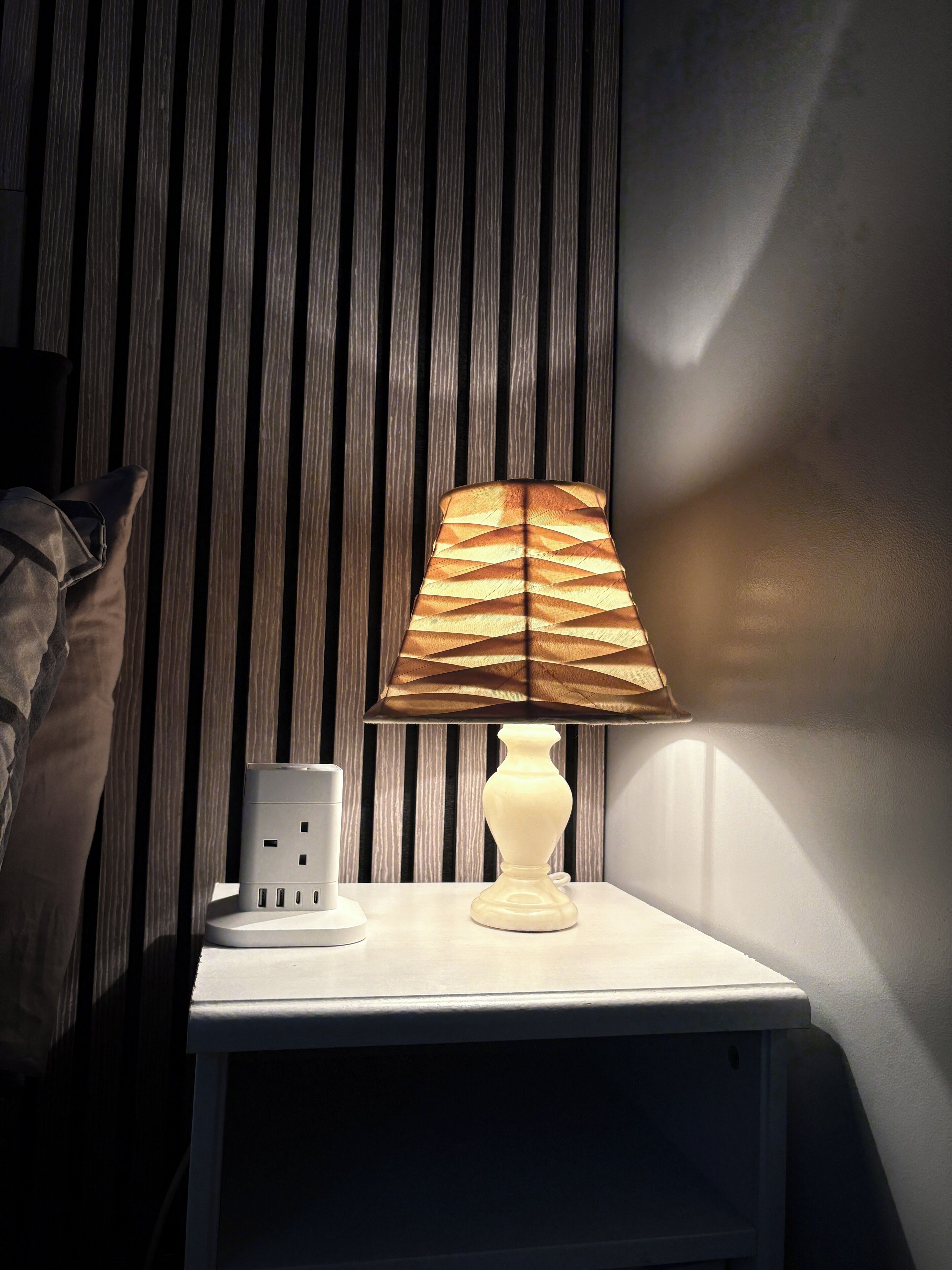 A bedside table with a lamp, a power strip, and a pillow against a striped wall.