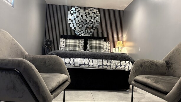 A modern bedroom with a bed, two armchairs, a wall-mounted mirror, and a bedside lamp. - The Garden Studio (Luton)