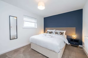 A bedroom with a bed, a bedside table, a nightstand, a mirror, and a window with blinds. - The Gordon | Vacant Nests | Heart of Aberdeen (Aberdeen)