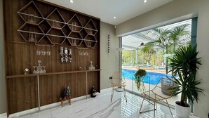 A modern interior with a wooden wall unit displaying glassware, a poolside seating area, and a large window offering a view of a swimming pool. - Bluebell House (London)