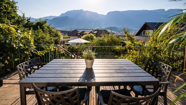 Een houten tafel met stoelen en een potplant, op een balkon met uitzicht op een bergachtig landschap.