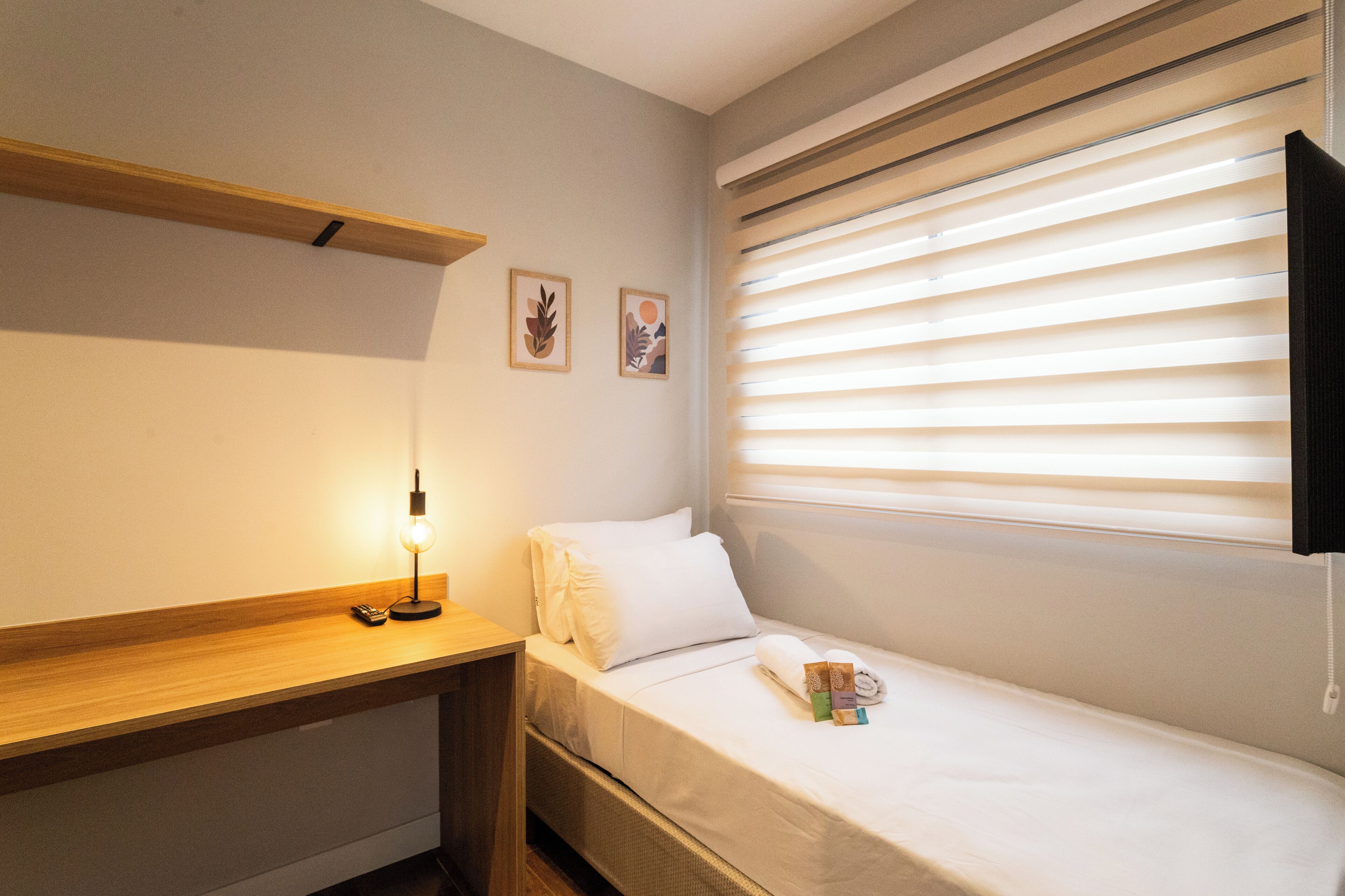 Un dormitorio pequeño y ordenado con una cama individual, una mesita de noche con lámpara, un estante de madera y una ventana con persianas.
