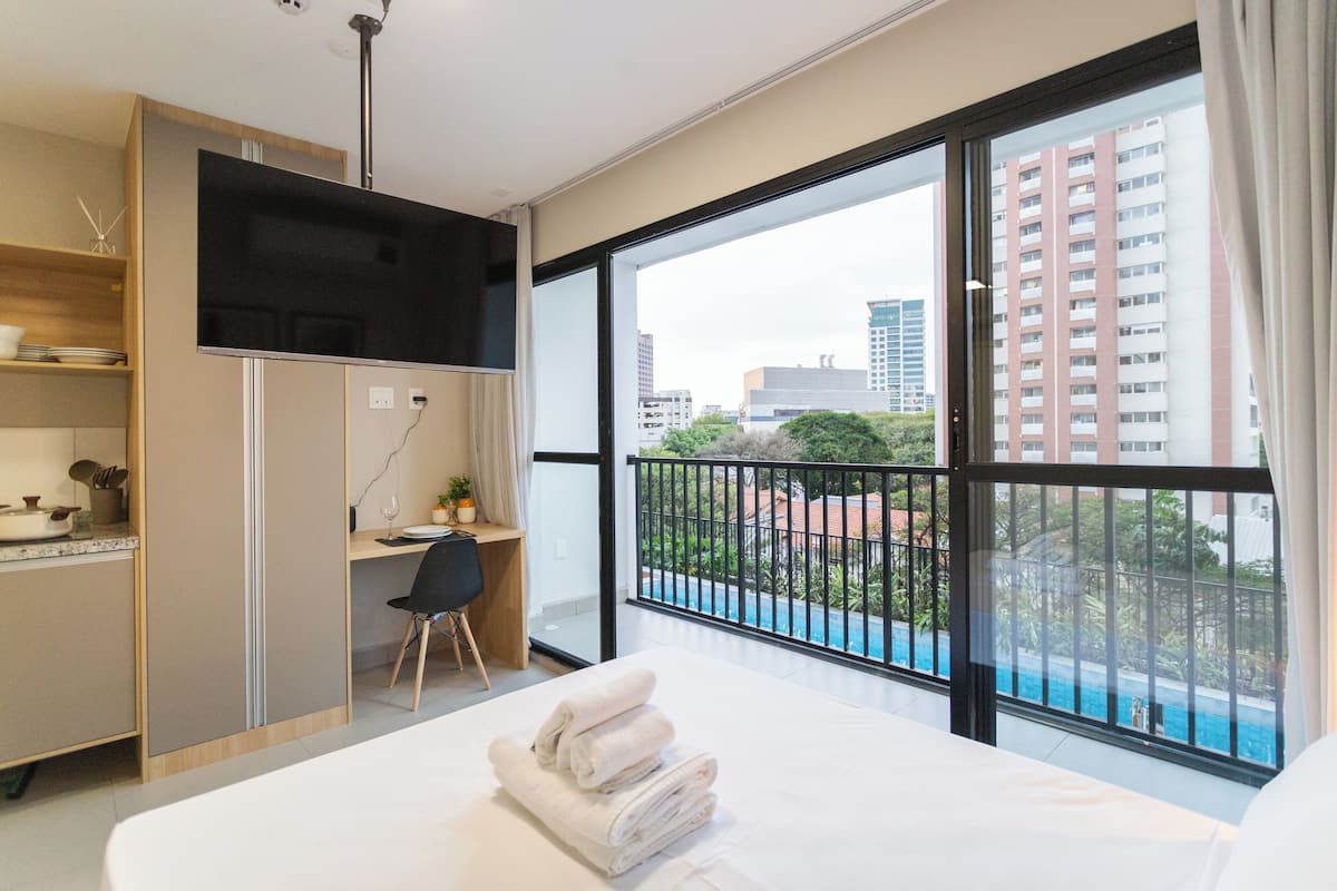 A modern hotel room with a large bed, a desk, and a view of a cityscape and a swimming pool.
