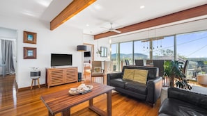 A modern living room with a flat-screen TV, wooden furniture, a black leather sofa, and a large window offering a view of the outdoors. - We’re thrilled to introduce this stunning new listing with The Lifestyle Group, Crescent Head. (Crescent Head)