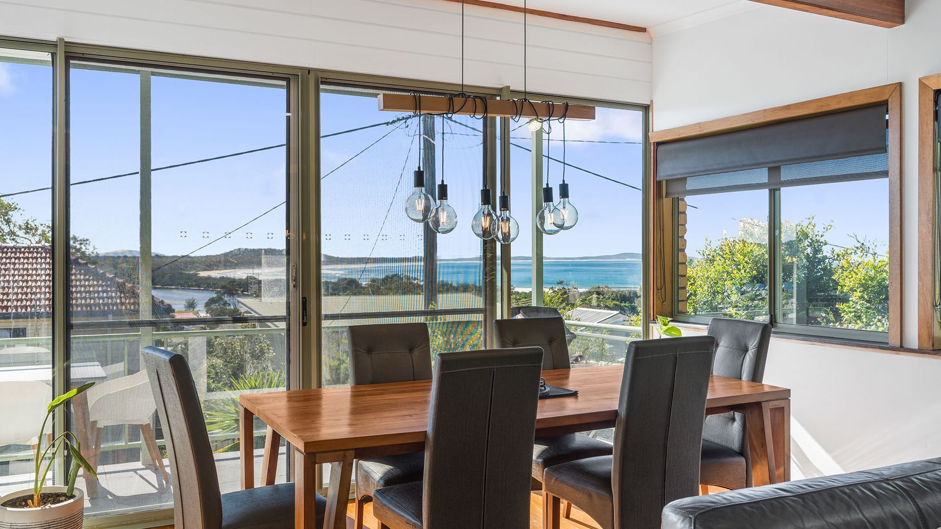 Un espace repas comprenant une table et des chaises, de grandes fenêtres offrant une vue sur la mer, et des luminaires suspendus.