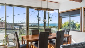 Un espace repas comprenant une table et des chaises, de grandes fenêtres offrant une vue sur la mer, et des luminaires suspendus.
