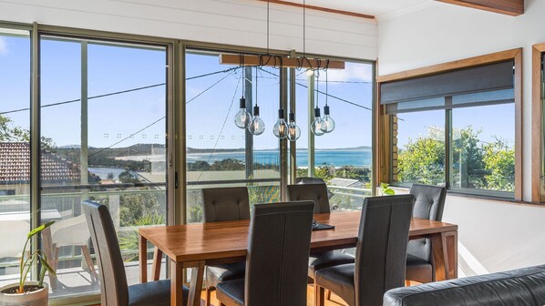 Een eethoek met een tafel en stoelen, grote ramen met uitzicht op zee en hangende lampen.