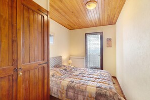 A bedroom with a wooden door, a bed with a patterned bedspread, a nightstand with a lamp, and a window with patterned curtains.