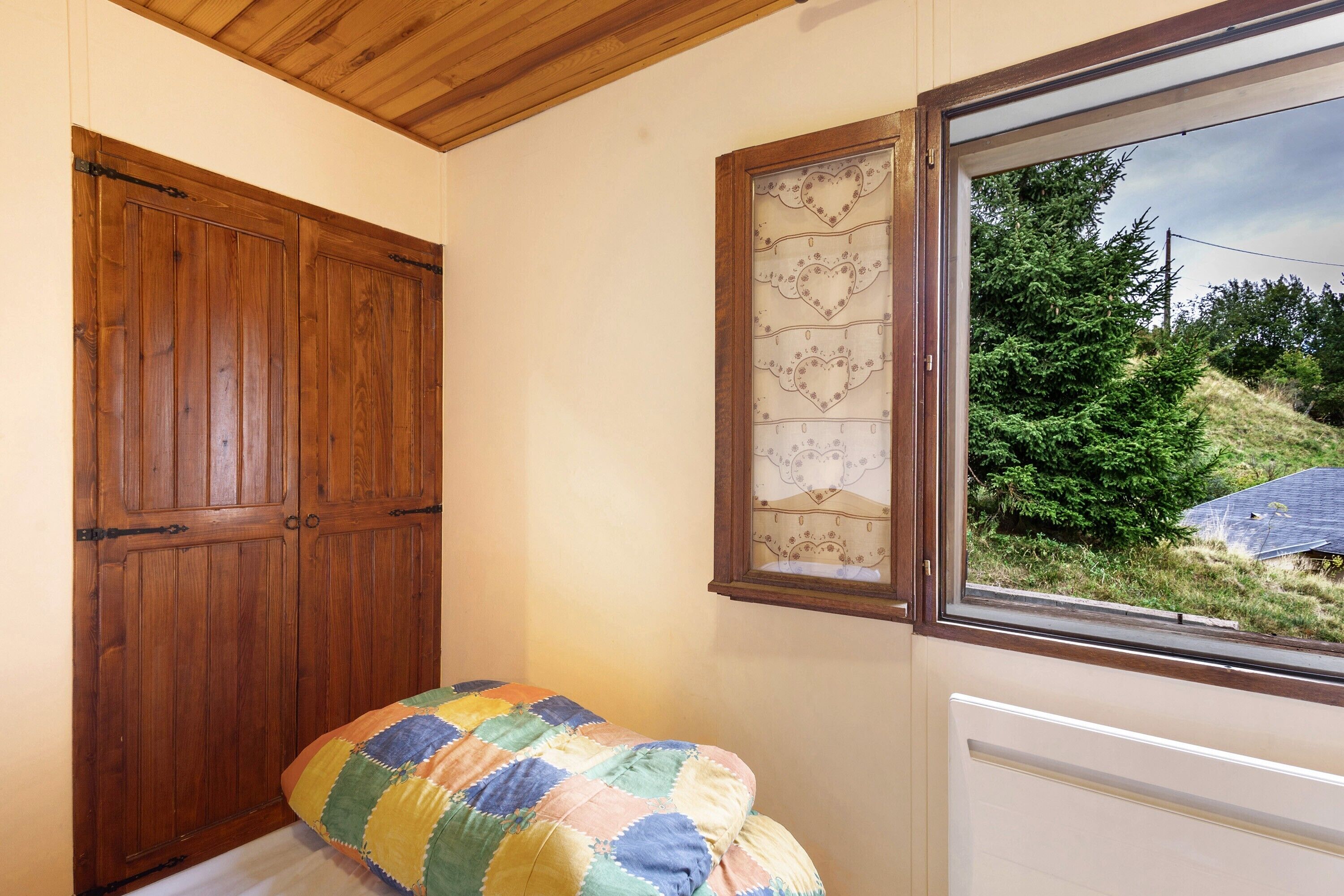 A room with a wooden wardrobe, a quilted bed, a window with a view of greenery, and a radiator.