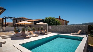 Une piscine entourée de chaises longues et de parasols, bordée de murs en pierre et offrant une vue sur les collines.
