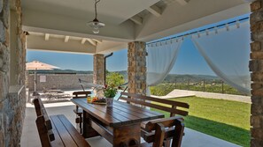 Une table en bois, des bancs et un pot de fleurs sont dispos és devant un mur de pierre, offrant une vue sur un paysage verdoyant et des collines lointaines.