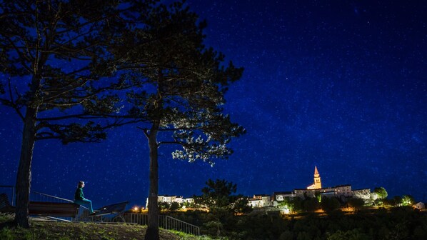 Une scène nocturne avec un ciel étoilé, une personne assise sur un banc et un bâtiment avec une tour en arrière-plan.