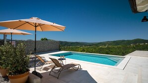 Un área de piscina con sillones de descanso y sombrillas, rodeada por un muro de piedra y con vistas a un paisaje pintoresco.