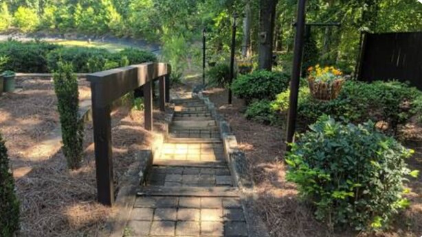 A stone pathway leading through a garden with a wooden fence on one side and a black fence on the other. - Wooded Oasis: Safe & Secluded 2BR BASEMENT APT, Full Bath Retreat (Stockbridge)