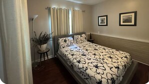 A bedroom with a bed, curtains, a potted plant, and two framed pictures on the wall. - Wooded Oasis: Safe & Secluded 2BR BASEMENT APT, Full Bath Retreat (Stockbridge)
