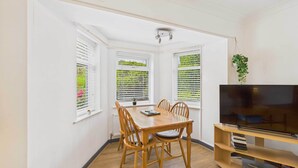 A dining area with a wooden table and chairs, a television on a wooden stand, and windows with blinds. - Host & Stay | LOOK Flat (Scarborough)