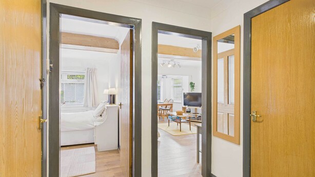 A hallway leading to a bedroom with a bed, bedside table, and lamp. There is a dining area with a table and chairs in the background. - Host & Stay | LOOK Flat (Scarborough)