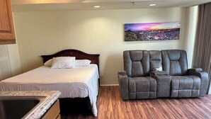 A hotel room with a bed, a reclining chair, a wooden floor, and a cityscape painting on the wall. - Stay steps from the sand in this private studio at the Imperial Resort. Walk to shops, dining & nightlife. Rooftop pool, hot tub, gym, and kitchen included! (Honolulu)