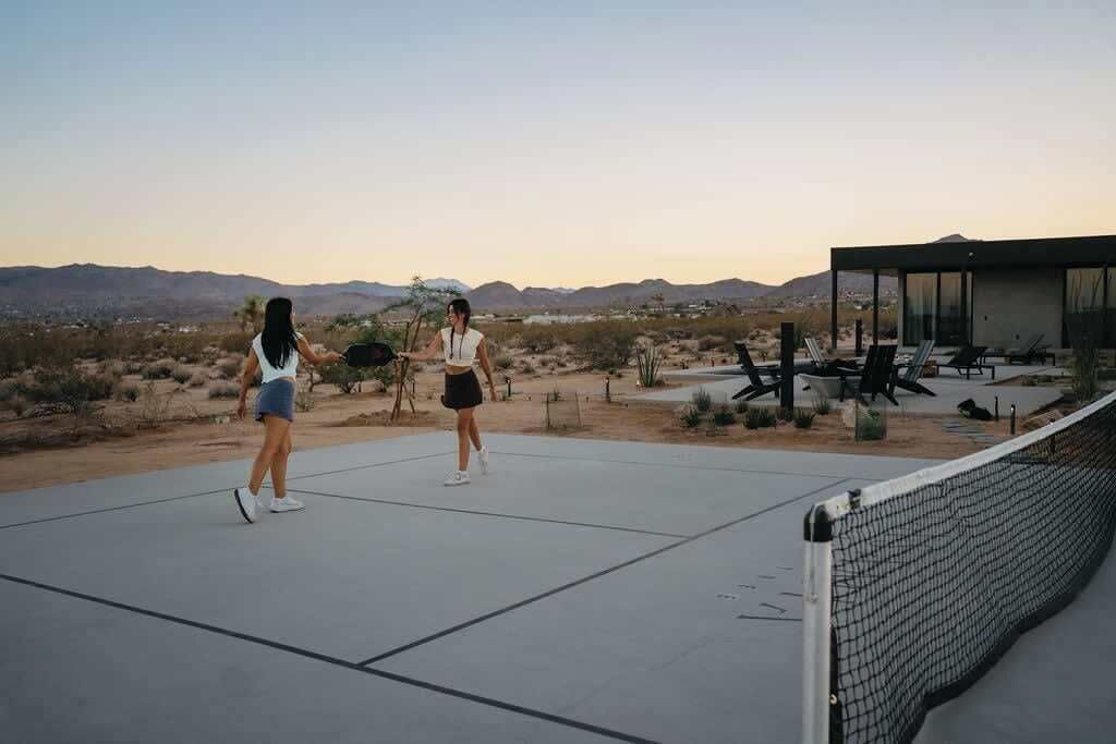 Una pista de tennis a l'aire lliure amb dues persones jugant, envoltada per un paisatge desèrtic i un edifici modern amb façana de vidre.