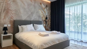Camera d'hotel moderna con un letto grande, un comodino, una lampada e vista su palme attraverso tende trasparenti.