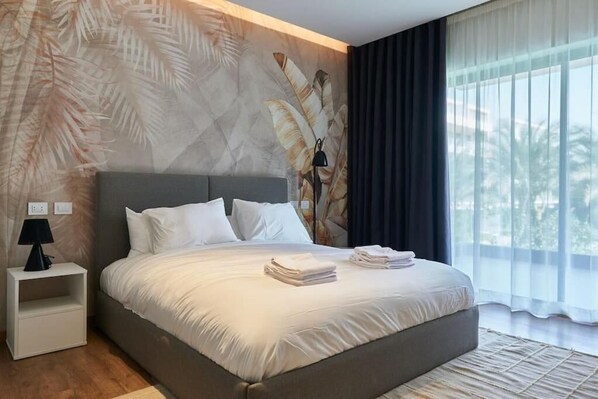 A modern hotel room with a large bed, a nightstand, a lamp, and a view of palm trees through sheer curtains.