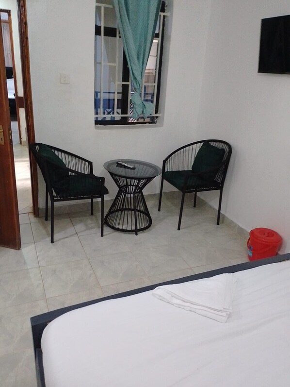 A room with a bed, two chairs, a small table, a window with curtains, and a red bucket on the floor.