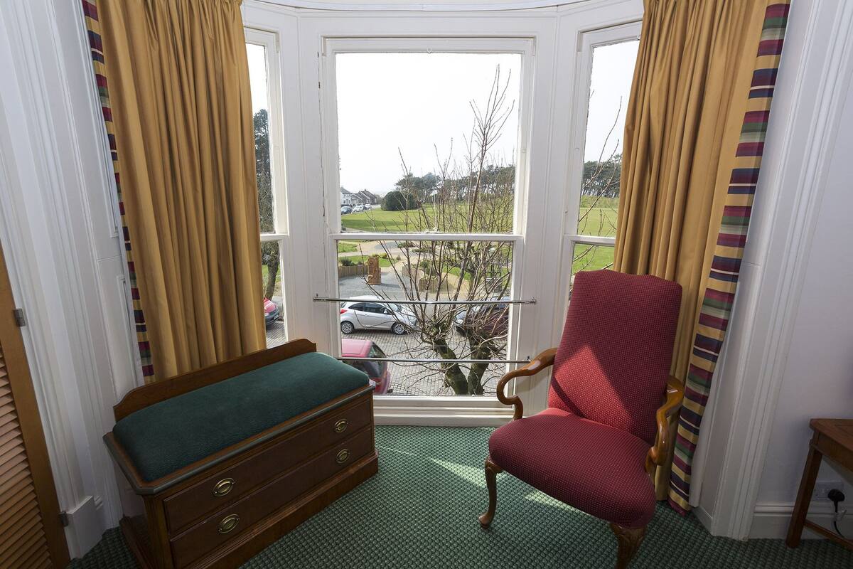 A corner room with a bay window, a wooden bench, a red armchair, and a view of a street with cars and greenery.