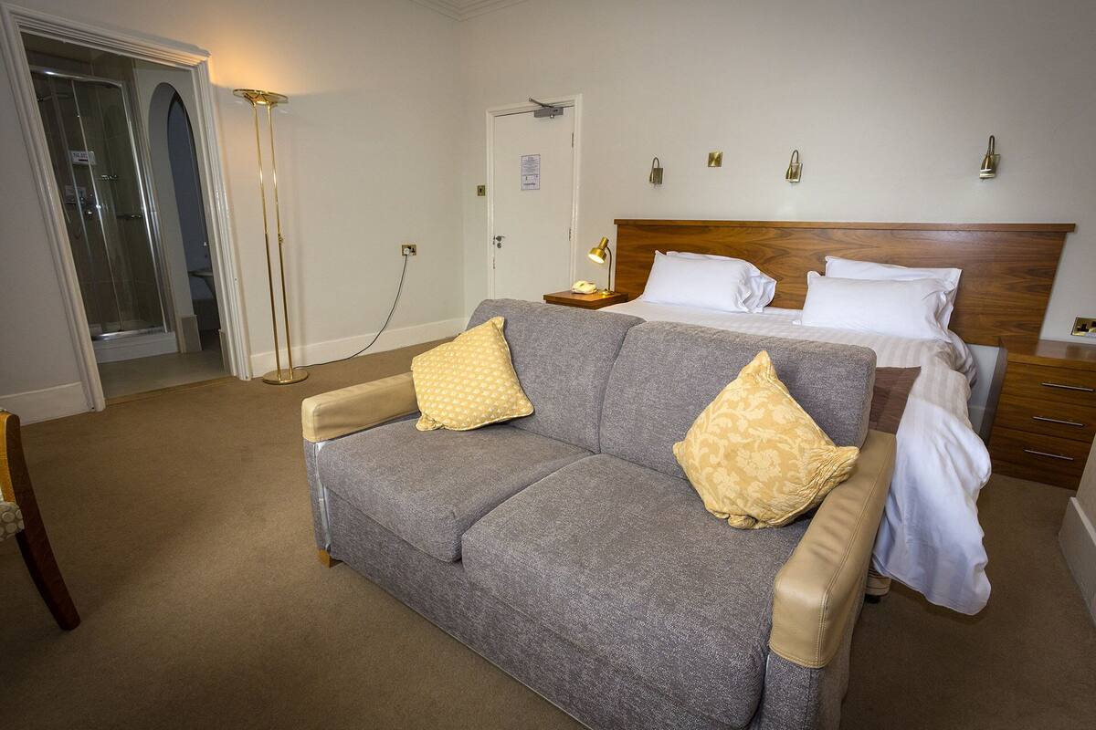 A hotel room with a grey sofa, two pillows, a wooden headboard, a bedside table with a lamp, and a bathroom visible through a glass door.