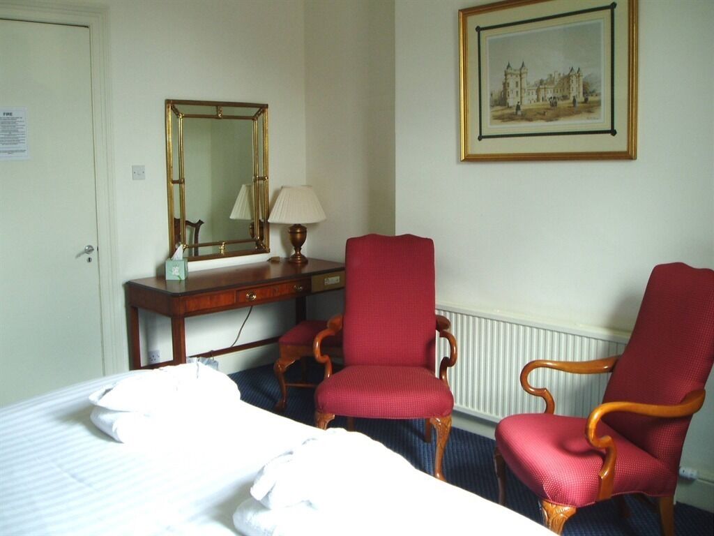 Una habitación de hotel con una cama, dos sillones tapizados en rojo, un escritorio de madera con una lámpara, un espejo y un cuadro en la pared.