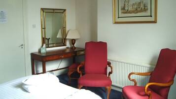 Una habitación de hotel con una cama, dos sillones tapizados en rojo, un escritorio de madera con una lámpara, un espejo y un cuadro en la pared.