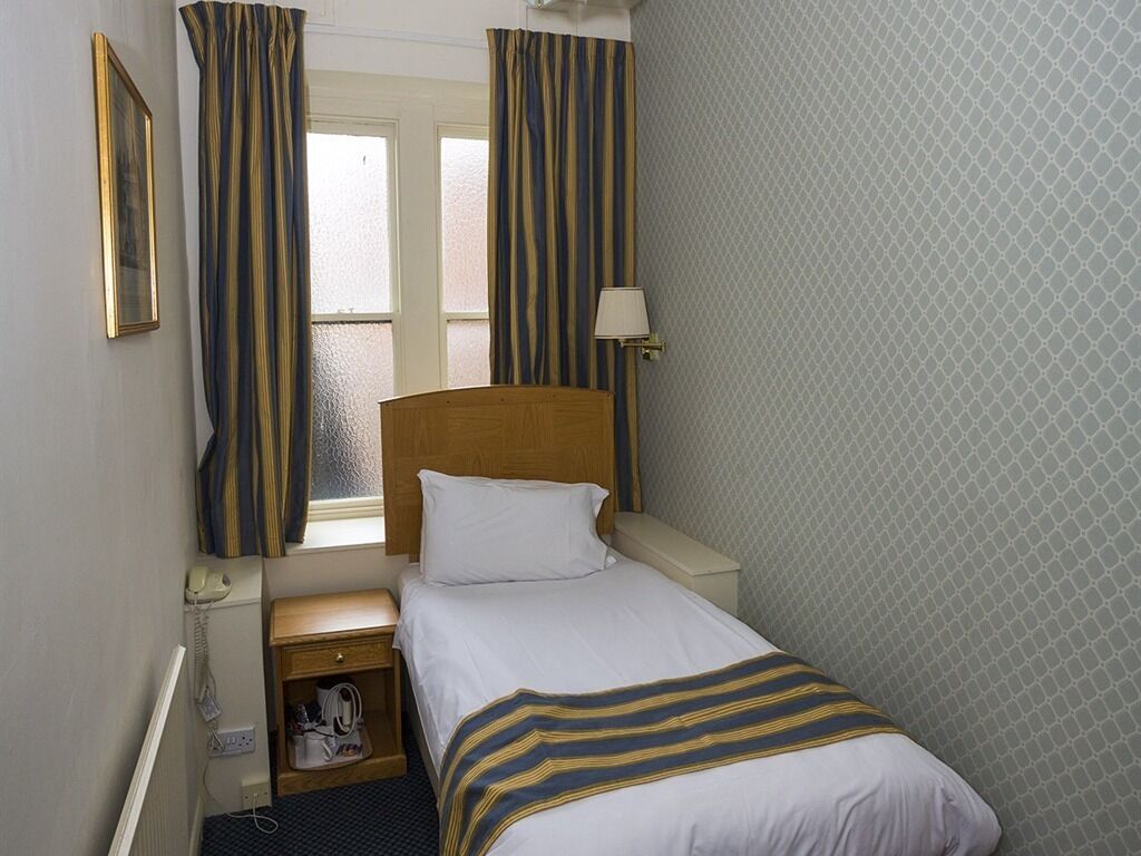 A hotel room with a single bed, a bedside table, a lamp, a telephone, and a window with curtains.