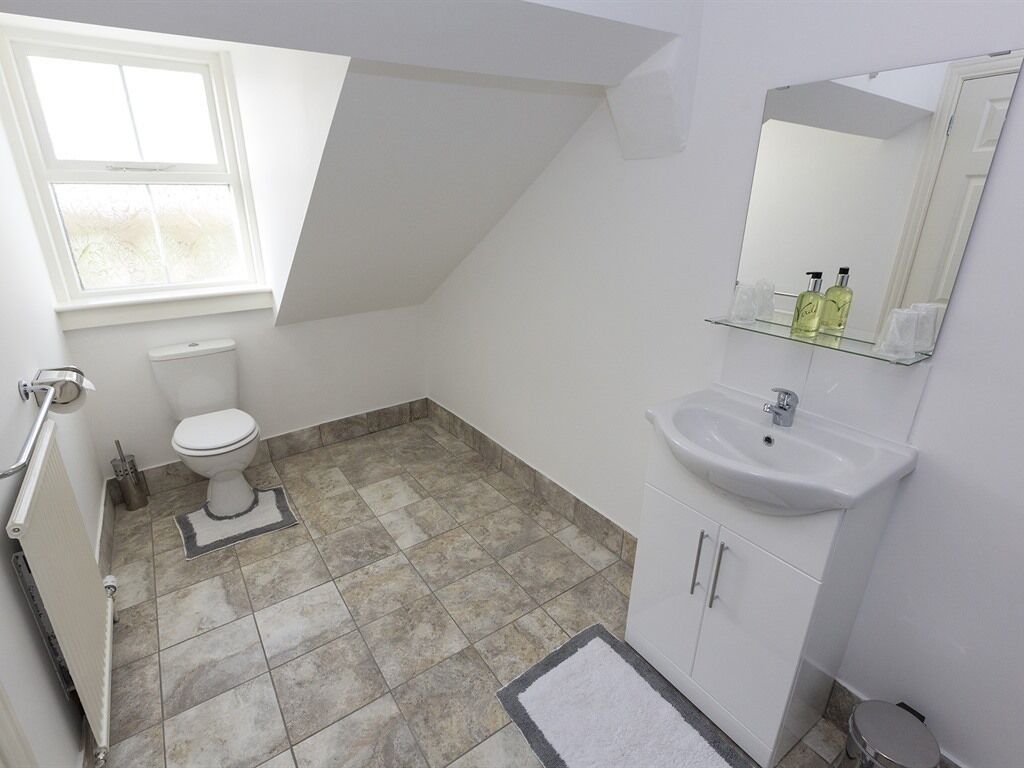 A small, well-lit bathroom with a toilet, sink, and mirror. It has a sloped ceiling and a window providing natural light.