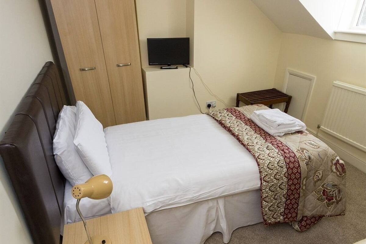 A bedroom with a bed, a bedside table with a lamp, a TV on a stand, a wardrobe, and a radiator.