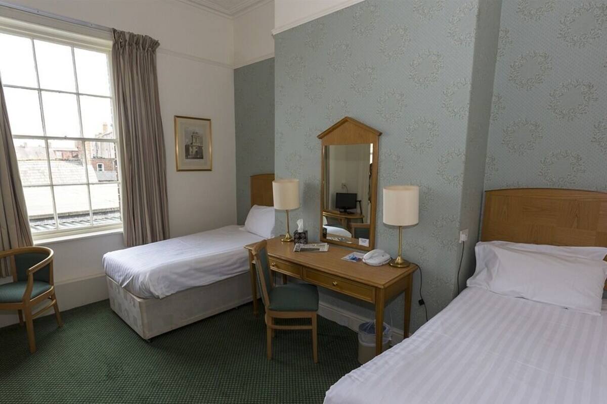 A hotel room with two single beds, a desk with a chair, a mirror, and a window with curtains.
