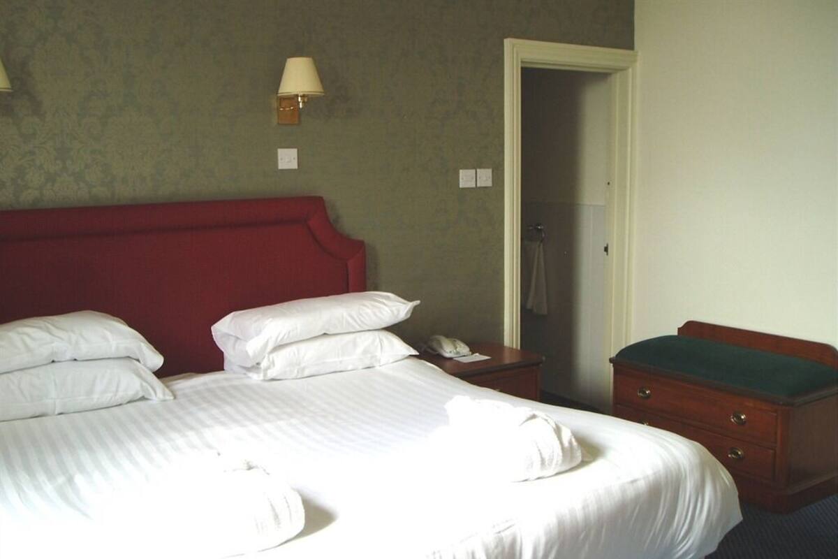 A neatly made bed with white linens, a wooden nightstand with a lamp, and a wooden dresser with a green cloth.